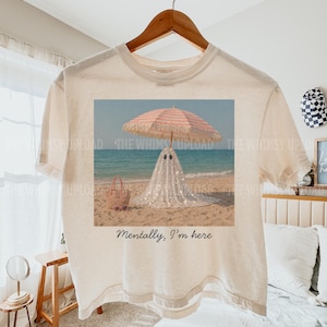 May include: Off-white t-shirt with a beach scene graphic. The graphic shows a ghost in a sparkly outfit under a pink and white striped parasol. The text on the shirt says "Mentally, I'm here."