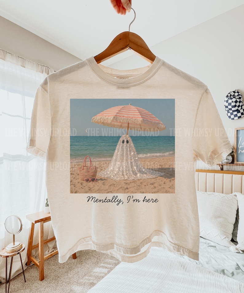 May include: Off-white t-shirt with a beach scene graphic. The graphic shows a ghost in a sparkly outfit under a pink and white striped parasol. The text on the shirt says "Mentally, I'm here."