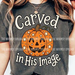 May include: A charcoal gray t-shirt featuring a graphic of an orange jack-o'-lantern pumpkin decorated with white stars and dots. The text "Carved in His Image" is printed in a cream-colored font above and below the pumpkin illustration.
