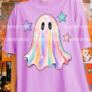 May include: A light purple t-shirt featuring a central graphic of a ghost with a multicolored iridescent finish, two black oval eyes, and small white star accents. Three solid-colored stars in purple, teal, and pink surround the ghost.