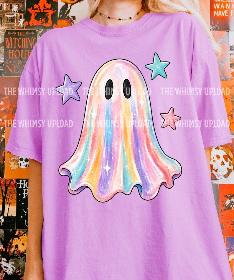 May include: A light purple t-shirt featuring a central graphic of a ghost with a multicolored iridescent finish, two black oval eyes, and small white star accents. Three solid-colored stars in purple, teal, and pink surround the ghost.