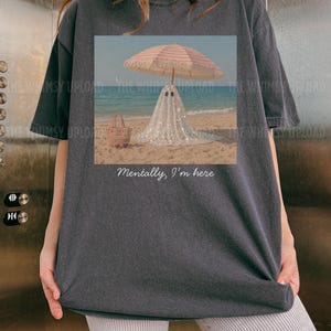 May include: Dark grey t-shirt with a beach scene print. A ghost figure under a striped parasol on the sand, with a tote bag and sunglasses. The text "Mentally, I'm here" is below the image. The sea and sky are visible in the background.