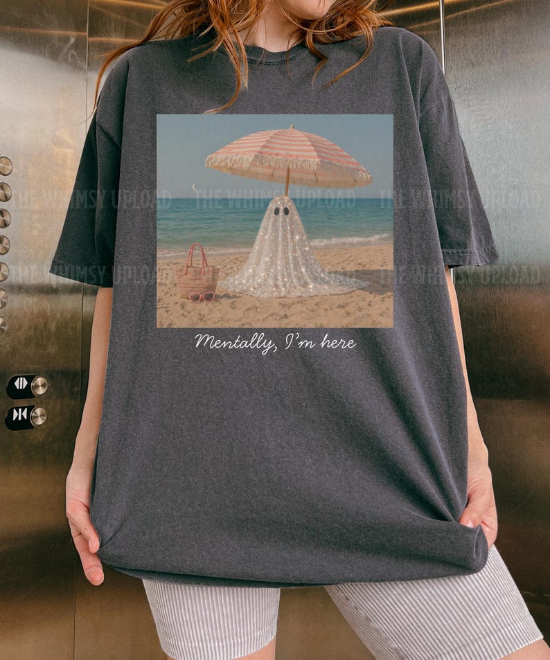 May include: Dark grey t-shirt with a beach scene print. A ghost figure under a striped parasol on the sand, with a tote bag and sunglasses. The text "Mentally, I'm here" is below the image. The sea and sky are visible in the background.