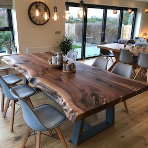 May include: A large rectangular dining table with a natural live edge wood slab top featuring dark brown grain patterns, supported by a sturdy black metal U-shaped base, surrounded by several light gray molded chairs with wooden legs.