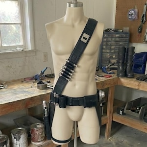 May include: A mannequin displays a black leather utility belt with silver bullet holders, a shoulder strap, and a holster. The mannequin is on a metal stand in a workshop setting.