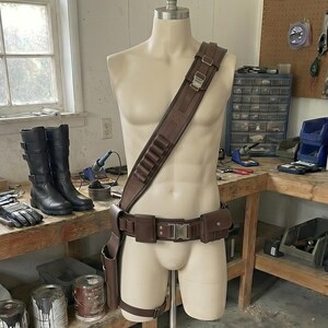 以下が含まれることがあります： マネキンに着せられた茶色の革のコスチュームセット。ショルダー ストラップ、ホルスター付きベルト、レッグ ストラップが含まれます。黒いブーツと手袋が近くのテーブルにあります。セットはワークショップ環境で表示されます。