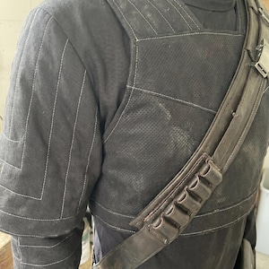 May include: Black costume with white stitching. The shoulder area features a quilted design. A brown leather strap with pouches is worn across the chest. The costume appears to be made of durable fabric.