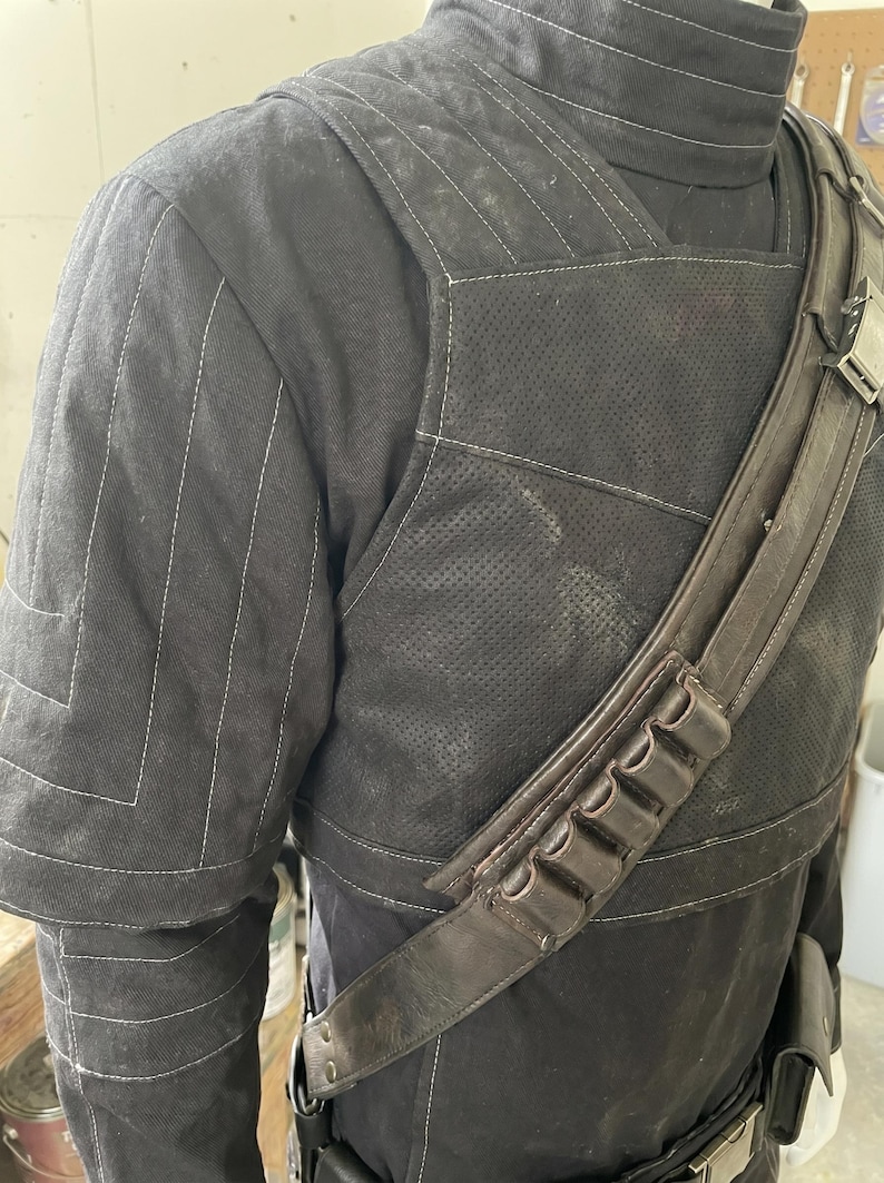May include: Black costume with white stitching. The shoulder area features a quilted design. A brown leather strap with pouches is worn across the chest. The costume appears to be made of durable fabric.