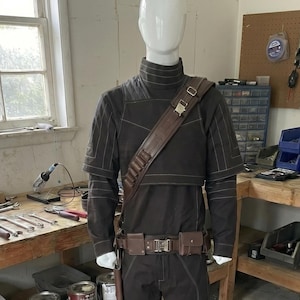 May include: A dark brown costume displayed on a mannequin. The outfit includes a long-sleeved top, pants, belt, and shoulder strap, all in matching shades. The costume also features a leg wrap and a holster. The mannequin is in a workshop setting.