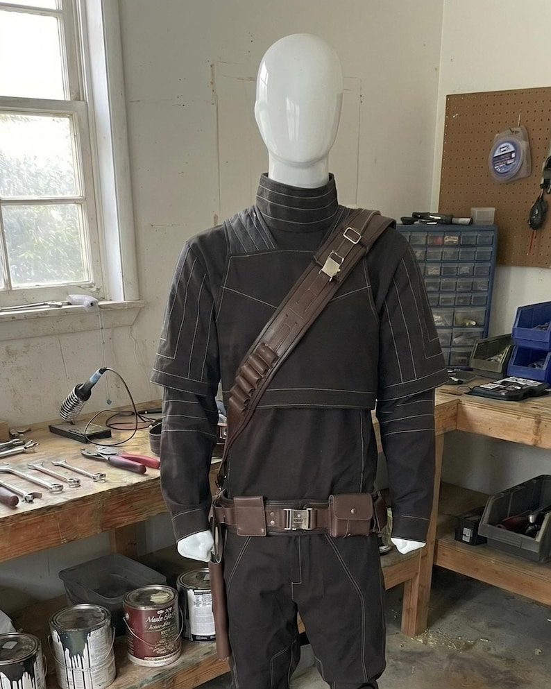May include: A dark brown costume displayed on a mannequin. The outfit includes a long-sleeved top, pants, belt, and shoulder strap, all in matching shades. The costume also features a leg wrap and a holster. The mannequin is in a workshop setting.