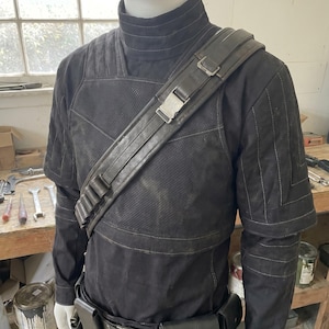 May include: Black costume with a high collar, shoulder strap, and belt with pouches. The costume has a textured appearance with stitched details. The shoulder strap features a metal buckle. The outfit is displayed on a mannequin.