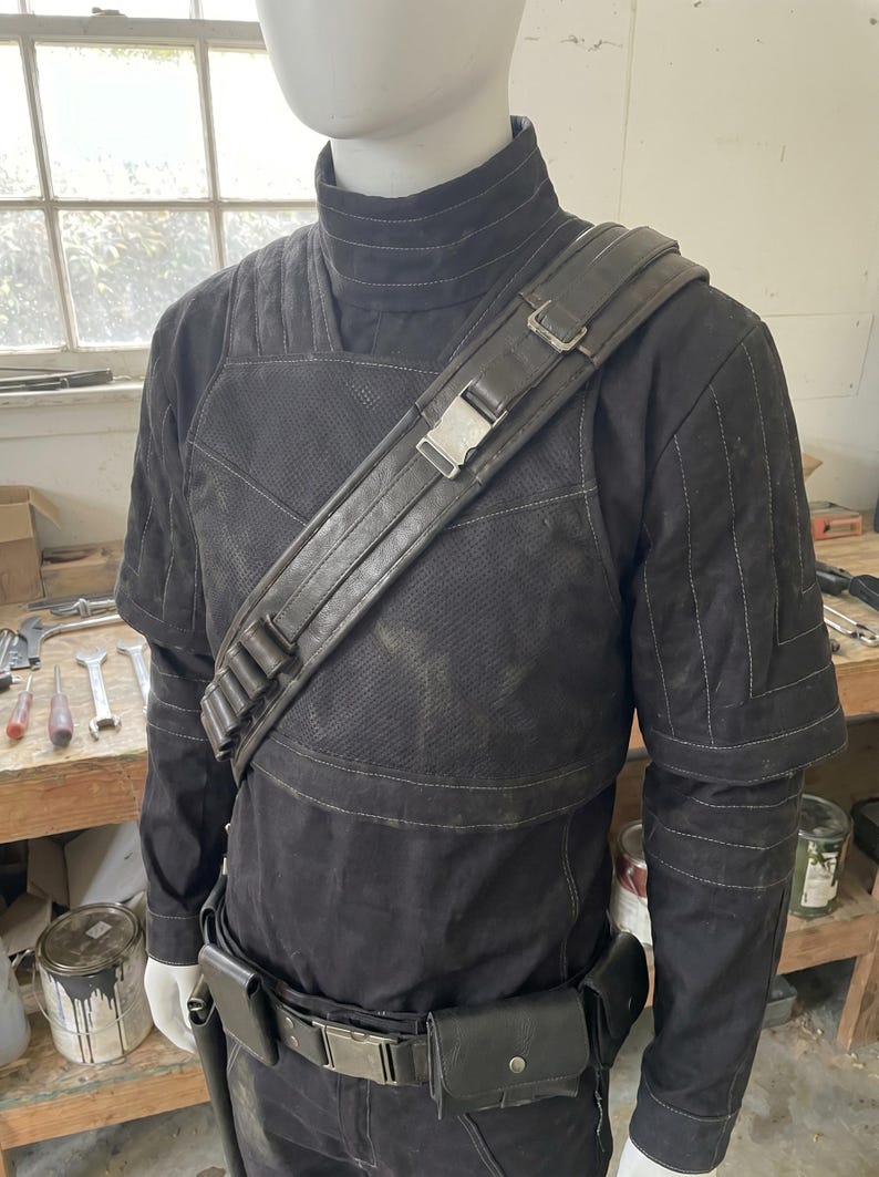 May include: Black costume with a high collar, shoulder strap, and belt with pouches. The costume has a textured appearance with stitched details. The shoulder strap features a metal buckle. The outfit is displayed on a mannequin.