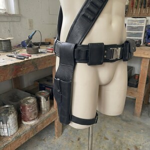 May include: Black leather gun holster and belt rig displayed on a mannequin. The rig includes a shoulder strap, belt with pouches, and a leg strap. The holster is designed to hold a handgun. The mannequin is in a workshop setting.