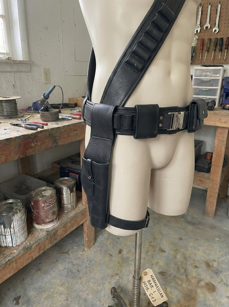May include: Black leather gun holster and belt rig displayed on a mannequin. The rig includes a shoulder strap, belt with pouches, and a leg strap. The holster is designed to hold a handgun. The mannequin is in a workshop setting.
