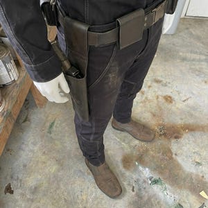May include: A person in a black jumpsuit with a leather belt and a gun holster. The holster contains a gun. The person is also wearing brown shoes and white gloves. The image is taken from a medium distance.
