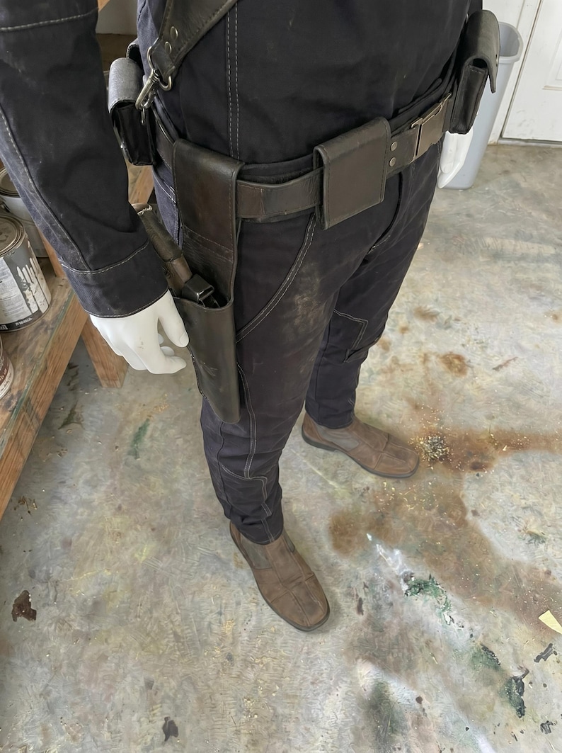 May include: A person in a black jumpsuit with a leather belt and a gun holster. The holster contains a gun. The person is also wearing brown shoes and white gloves. The image is taken from a medium distance.