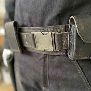 May include: A dark brown leather belt with a large, silver-coloured buckle. The belt has two attached pouches and is worn over dark denim trousers. The buckle is rectangular and has a vintage, worn appearance. The belt is a practical accessory.