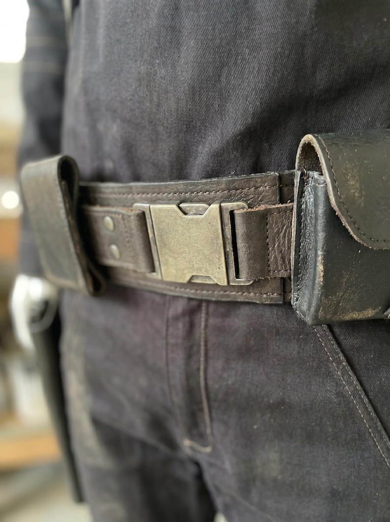 May include: A dark brown leather belt with a large, silver-coloured buckle. The belt has two attached pouches and is worn over dark denim trousers. The buckle is rectangular and has a vintage, worn appearance. The belt is a practical accessory.