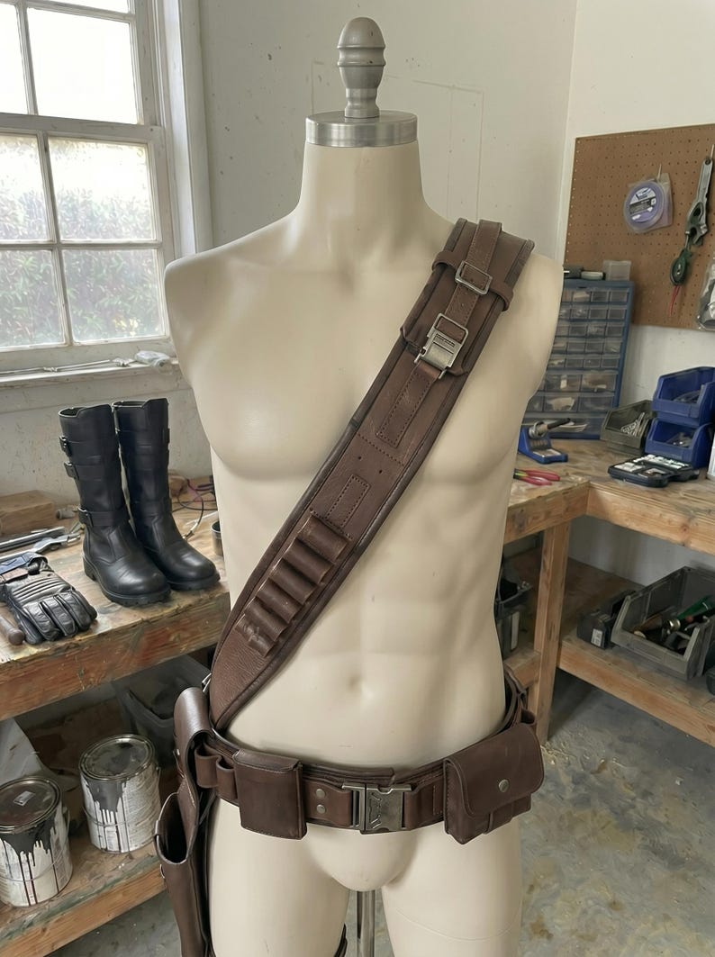 以下が含まれることがあります： マネキンの上に展示された茶色の革製ユーティリティベルトとショルダーストラップのセット。ベルトには複数のポーチと装飾的なバックルが付いています。ショルダーストラップにはカートリッジホルダーと調節可能なバックルが付いています。背景には黒いブーツと手袋が見えます。