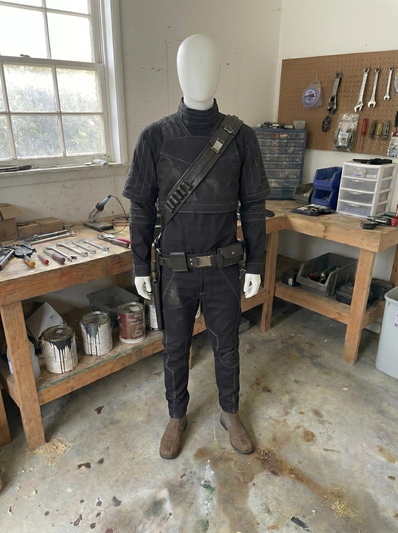 May include: A black costume with a high collar, shoulder strap, and belt, displayed on a mannequin. The outfit includes a long-sleeved top, pants, and boots. The mannequin is in a workshop setting with tools and paint cans visible.