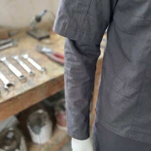 Puede incluir: Una camisa de trabajo gris oscuro con mangas cortas y detalles de costura visibles. La camisa es parte de un uniforme de trabajo, y la imagen muestra una parte de la manga y la parte inferior de la camisa.