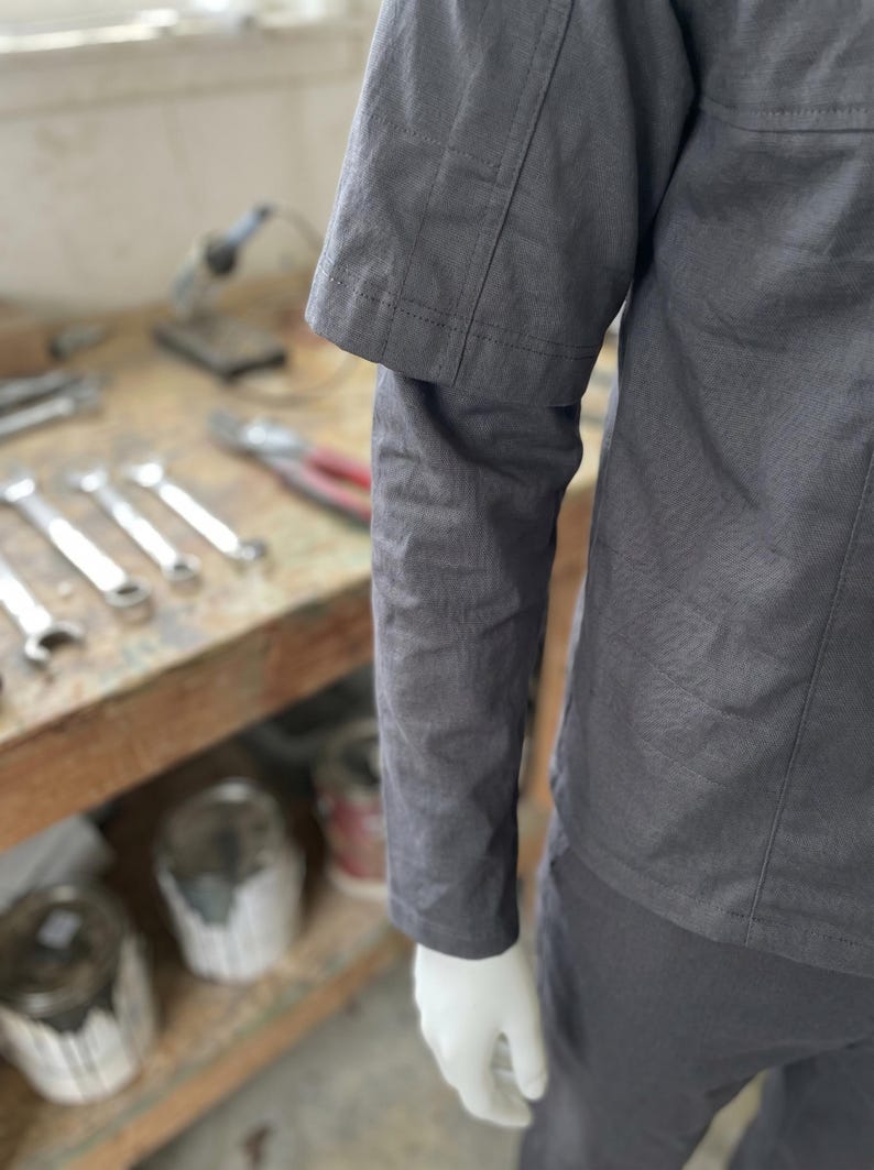Puede incluir: Una camisa de trabajo gris oscuro con mangas cortas y detalles de costura visibles. La camisa es parte de un uniforme de trabajo, y la imagen muestra una parte de la manga y la parte inferior de la camisa.