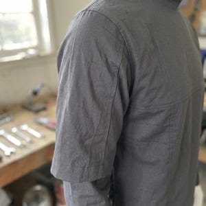 Puede incluir: Una camisa gris carb&oacute;n de manga corta con cuello alto. La camisa tiene un dise&ntilde;o minimalista con costuras visibles y un ajuste estructurado. La tela parece ser ligera, posiblemente una mezcla de lino o algod&oacute;n, adecuada para climas c&aacute;lidos.