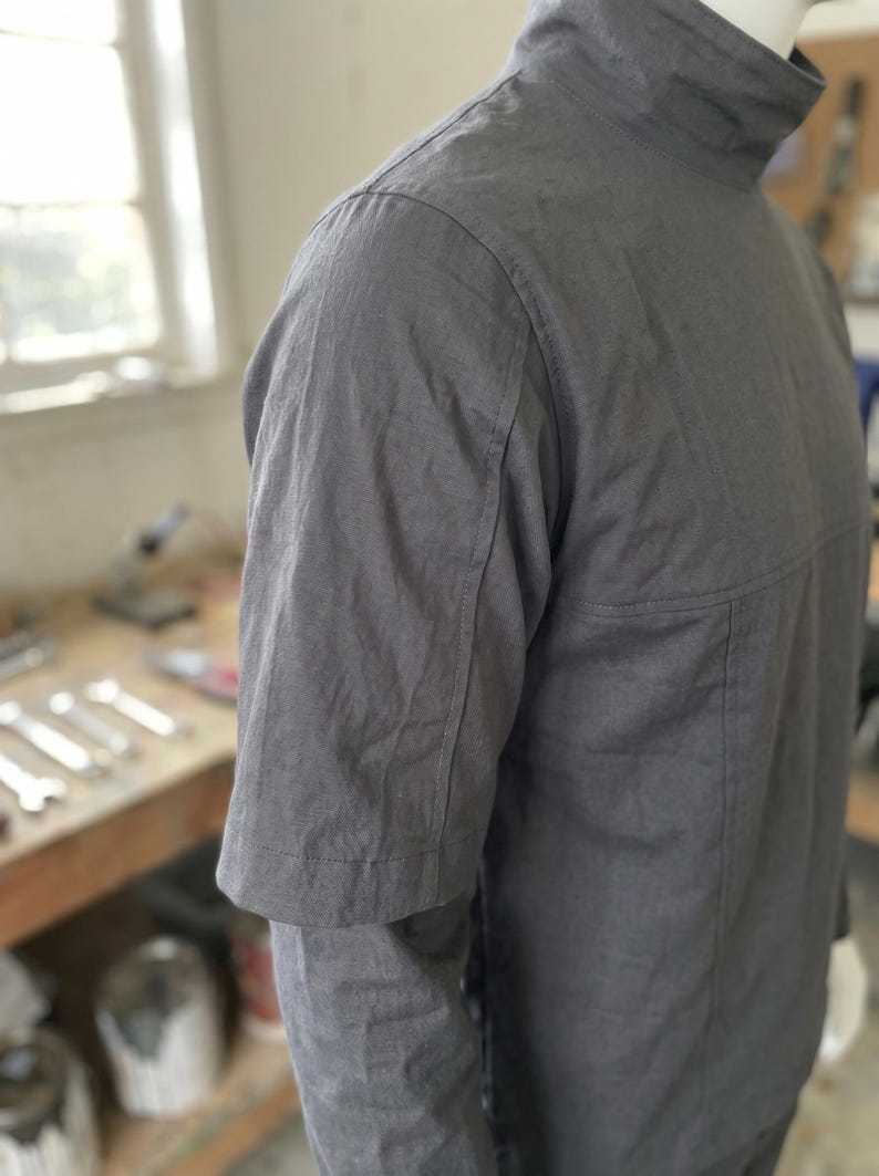 Puede incluir: Una camisa gris carb&oacute;n de manga corta con cuello alto. La camisa tiene un dise&ntilde;o minimalista con costuras visibles y un ajuste estructurado. La tela parece ser ligera, posiblemente una mezcla de lino o algod&oacute;n, adecuada para climas c&aacute;lidos.
