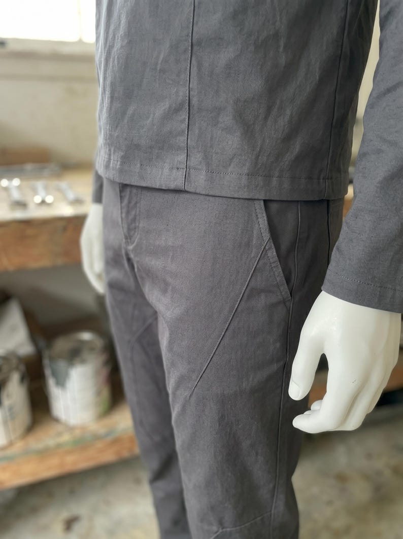 Puede incluir: Un conjunto gris de dos piezas sobre un maniqu&iacute;. La parte superior es una chaqueta de manga larga con costuras verticales. Los pantalones son grises con detalles de bolsillo en &aacute;ngulo. La mano del maniqu&iacute; es visible.