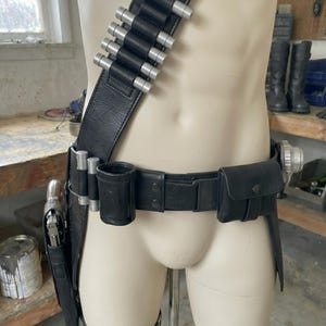 May include: Black leather utility belt with silver-tipped cartridge holders, a holster, and pouches. The belt is displayed on a mannequin. The belt includes a thigh strap and a shoulder strap.