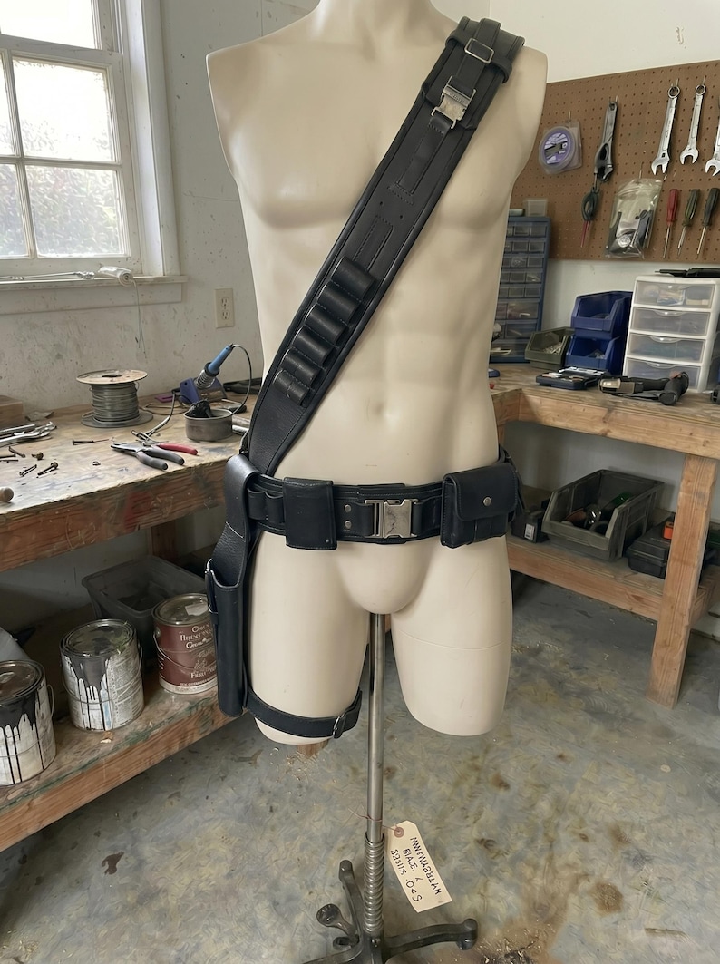 May include: Black leather utility belt and shoulder strap set displayed on a mannequin. The set includes a shoulder strap with cartridge holders, a belt with pouches, and a holster. The mannequin is on a metal stand.