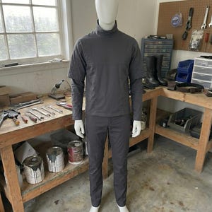 Puede incluir: Maniqu&iacute; con un atuendo gris oscuro, que incluye una parte superior de manga larga con cuello alto y pantalones a juego. El maniqu&iacute; est&aacute; en un taller, con herramientas y botes de pintura visibles en el fondo.