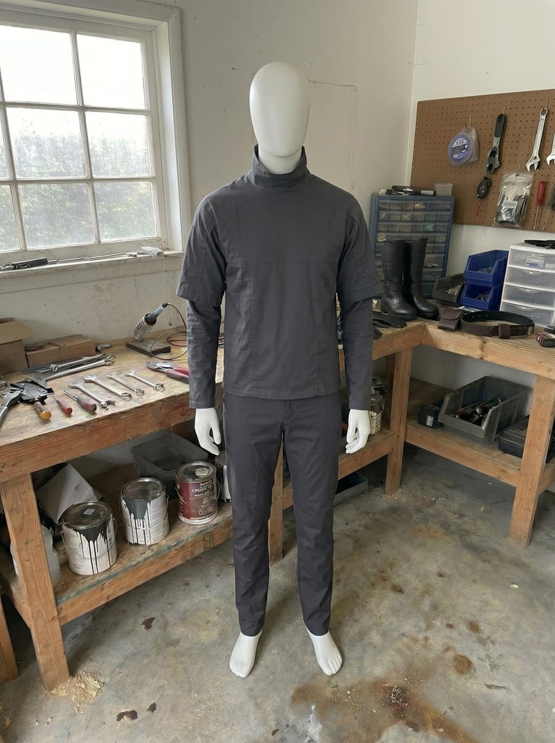 Puede incluir: Maniqu&iacute; con un atuendo gris oscuro, que incluye una parte superior de manga larga con cuello alto y pantalones a juego. El maniqu&iacute; est&aacute; en un taller, con herramientas y botes de pintura visibles en el fondo.