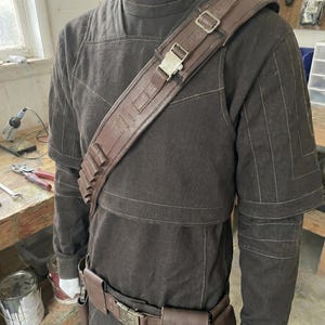 Puede incluir: Un disfraz marr&oacute;n con cuello alto, bandolera y cintur&oacute;n. El disfraz presenta una bandolera de cuero marr&oacute;n con hebilla met&aacute;lica, un cintur&oacute;n con bolsillos y una serie de porta-balas. El atuendo se exhibe en un maniqu&iacute;.