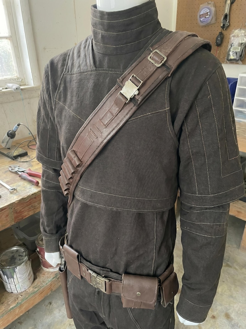 Puede incluir: Un disfraz marr&oacute;n con cuello alto, bandolera y cintur&oacute;n. El disfraz presenta una bandolera de cuero marr&oacute;n con hebilla met&aacute;lica, un cintur&oacute;n con bolsillos y una serie de porta-balas. El atuendo se exhibe en un maniqu&iacute;.