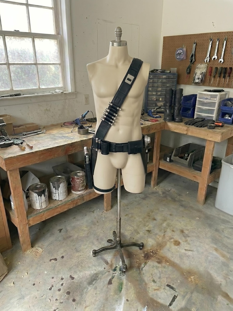 May include: A mannequin displays a black leather utility belt with silver bullet holders, a shoulder strap, and a holster. The mannequin is on a metal stand in a workshop setting.