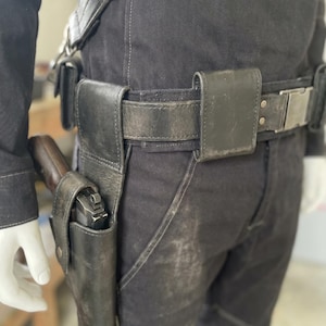 May include: Black leather gun holster and belt set. The belt has a silver buckle and several pouches. A handgun is visible in the holster. The set is worn on black trousers.