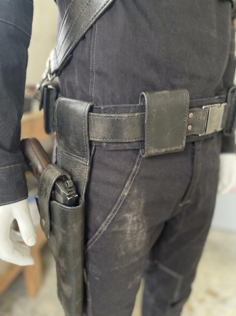 May include: Black leather gun holster and belt set. The belt has a silver buckle and several pouches. A handgun is visible in the holster. The set is worn on black trousers.