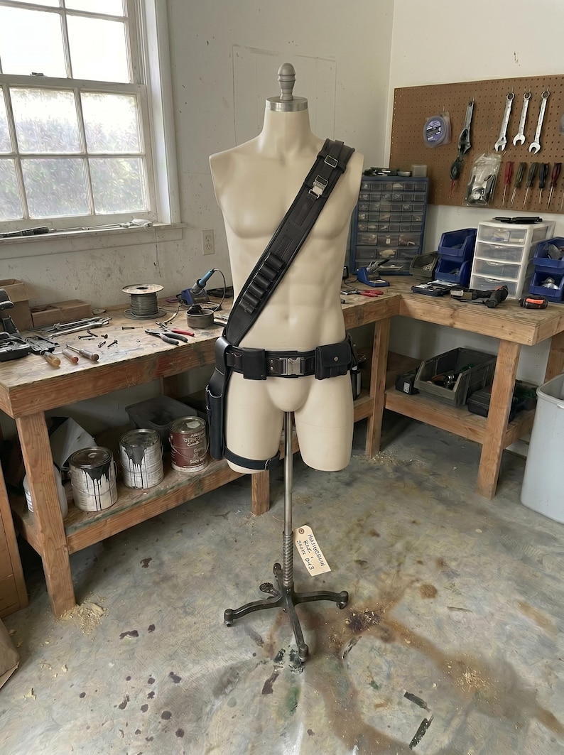 May include: A mannequin displays a black leather utility belt with pouches, a shoulder strap with cartridge holders, and a leg strap. The mannequin is in a workshop setting with tools and workbenches.