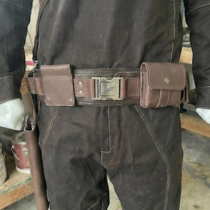 Puede incluir: Cintur&oacute;n utilitario marr&oacute;n con una hebilla grande y dos peque&ntilde;os bolsillos. El cintur&oacute;n es de cuero y tiene una hebilla rectangular. El cintur&oacute;n se lleva sobre pantalones marrones y una chaqueta marr&oacute;n.