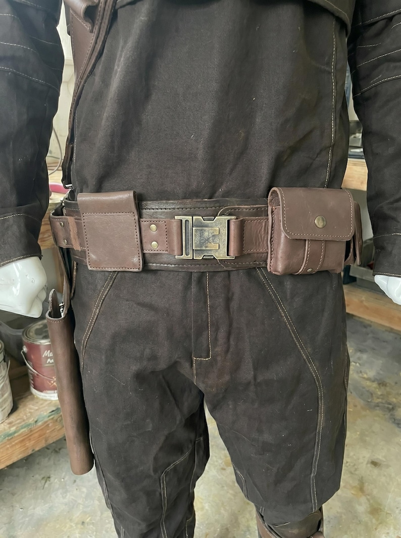 Puede incluir: Cintur&oacute;n utilitario marr&oacute;n con una hebilla grande y dos peque&ntilde;os bolsillos. El cintur&oacute;n es de cuero y tiene una hebilla rectangular. El cintur&oacute;n se lleva sobre pantalones marrones y una chaqueta marr&oacute;n.