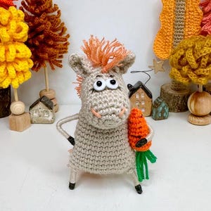 May include: A handmade crochet horse figurine in beige with orange hair and big eyes, holding a carrot. The horse is surrounded by small decorative trees in shades of orange and yellow, and miniature houses.