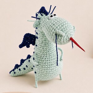 May include: A crocheted dragon toy in light blue with dark blue accents. The dragon has a red tongue, dark blue wings, and a row of dark blue spikes down its back. The toy is handmade and has a whimsical design.