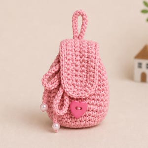 May include: A miniature, pink crocheted backpack. It features a flap secured by a heart-shaped button and a loop handle. The drawstring closure is adorned with two pearl beads. This handmade bag is a delightful accessory.