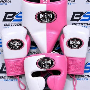 Puede incluir: Un conjunto de equipo de boxeo que incluye una coquilla protectora blanca y rosa, un casco a juego y dos guantes de boxeo. Cada pieza muestra un parche ovalado negro con el texto blanco NO BOXING NO LIFE. El equipo es de material liso.