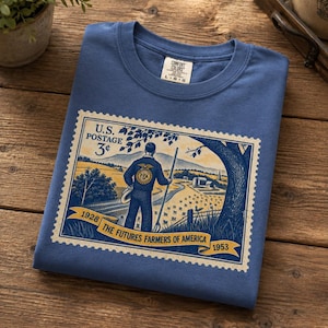 May include: A blue crew neck t-shirt featuring a graphic print of a vintage U.S. postage stamp. The design depicts a person standing in a field with the text U.S. POSTAGE 3 cents, 1928, THE FUTURES FARMERS OF AMERICA, and 1953. The shirt has a Comfort Colors label.