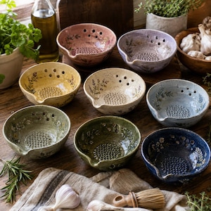 Puede incluir: Una colección de ocho cuencos de cerámica en varios colores, incluyendo rosa, morado, amarillo, crema, azul, verde y azul oscuro. Cada cuenco presenta una base ralladora texturizada, un pequeño pico vertedor y delicados motivos florales interiores.