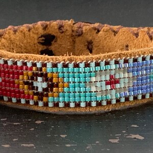 May include: A handcrafted leather bracelet with a beaded design. The bracelet features a tan leather band with a colorful pattern of red, blue, turquoise, and white beads. The beaded section is approximately 1 inch wide.