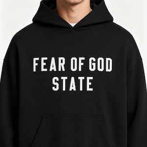 May include: Black hoodie with the text "FEAR OF GOD STATE" in white block letters. The hoodie has a front pocket and a hood. The person in the image is wearing a white undershirt.
