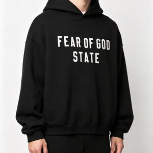 May include: Black hooded sweatshirt with the text "FEAR OF GOD STATE" in white block letters. The sweatshirt has long sleeves and a relaxed fit. The garment is made of a soft material.
